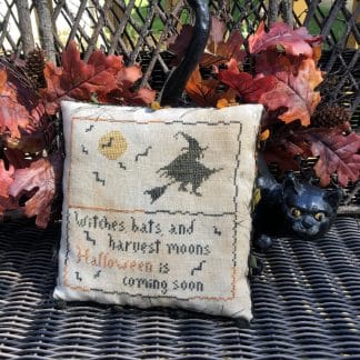Photo of finished Coming Soon pillow tuck by Falling Star Primitives, sitting on wicker bench against leaves and ceramic black cat