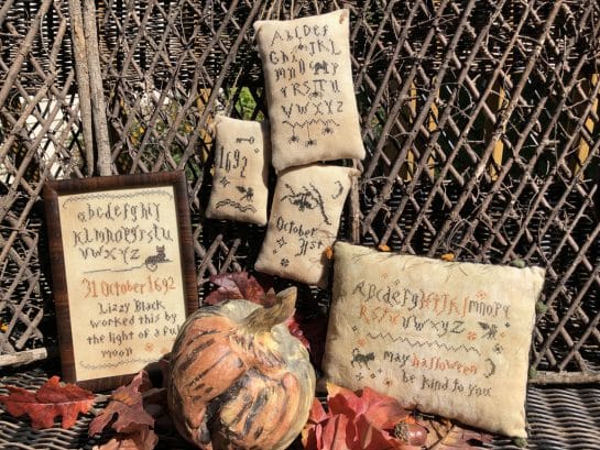 Photo of finished Wicked Little Samplers set by Falling Star Primitives, displayed against a wood screen with Autumn leaves and other Fall seasons decor
