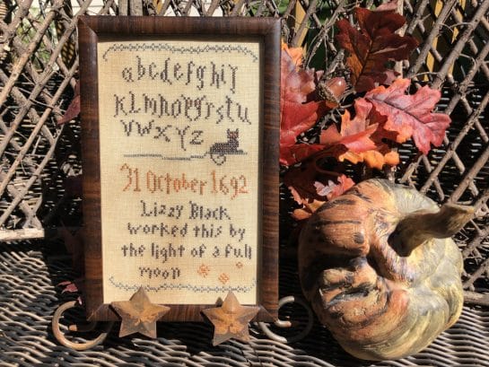 Photo of finished sampler from Wicked Little Samplers set by Falling Star Primitives, displayed against a wood screen with Autumn leaves and other Fall seasons decor