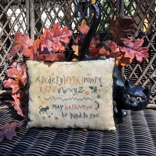 Photo of finished pillow tuck from Wicked Little Samplers set by Falling Star Primitives, displayed against a wood screen with Autumn leaves and other Fall seasons decor