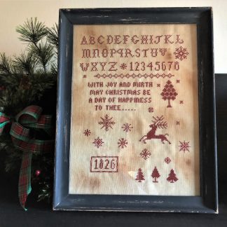 Photo of finished sampler from Joy & Mirth Redwork Sampler by Falling Star Primitives, displayed on top of evergreen branches on shelf