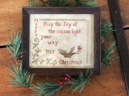 Photo of finished sampler from Light Your Way Sampler by Falling Star Primitives, displayed on top of evergreen branches