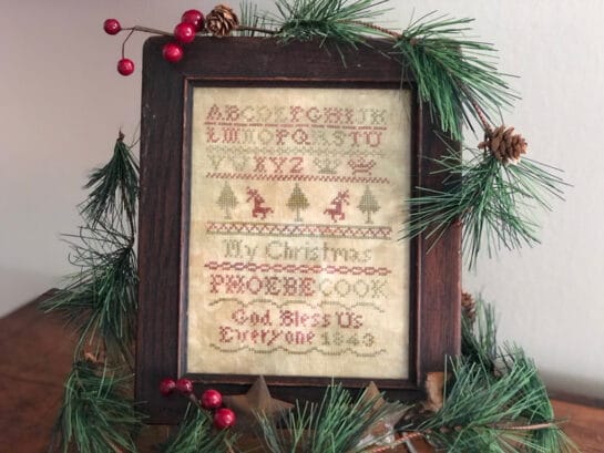Photo of finished sampler from Phoebes Christmas by Falling Star Primitives, displayed on top of evergreen branches on shelf
